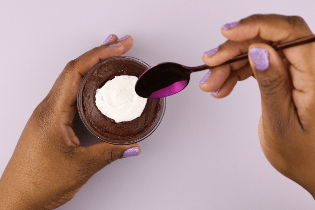 Hands with purple glittery nail polish holding a cake jar of chocolate dessert topped with a dollop of cream, with a purple spoon poised to take a scoop.