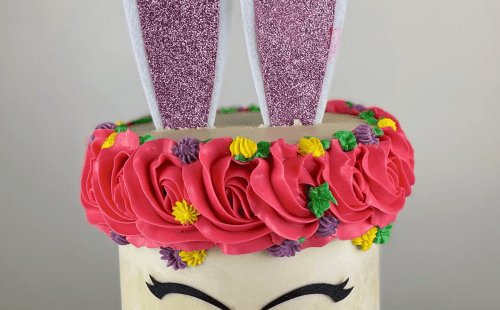 A cake designed to look like a bunny with pink ears, a heart nose, closed-eye lashes, and a floral crown.